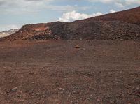 a lone horse standing in the dirt area with a hill behind it, with mountains behind it