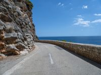 a curved mountain road that curves away from the ocean and next to the wall is a rock cliff