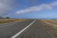 a long straight asphalt road near the ocean on a sunny day with a dark cloud overhead