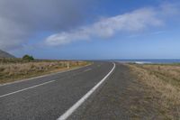 a long straight asphalt road near the ocean on a sunny day with a dark cloud overhead