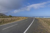 a long straight asphalt road near the ocean on a sunny day with a dark cloud overhead