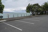 a parking lot is lined with trees next to the ocean and a bench sitting at the edge of it