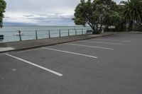 a parking lot is lined with trees next to the ocean and a bench sitting at the edge of it