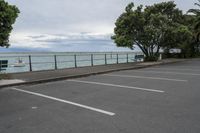 a parking lot is lined with trees next to the ocean and a bench sitting at the edge of it