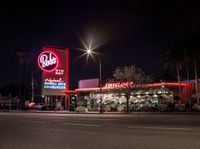 a red lit diner next to a palm tree lined street at night time with cars parked on side