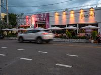 an old car drives past a restaurant at dusk in a city street with white tables with umbrellas