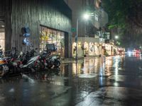 multiple motorcycles are parked along a wet street at night with shops and cars visible in the distance