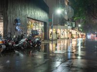 multiple motorcycles are parked along a wet street at night with shops and cars visible in the distance