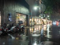 multiple motorcycles are parked along a wet street at night with shops and cars visible in the distance