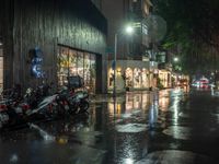 multiple motorcycles are parked along a wet street at night with shops and cars visible in the distance