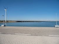 a brick street next to water under a blue sky with wind turbines in the distance