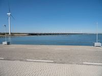 a brick street next to water under a blue sky with wind turbines in the distance