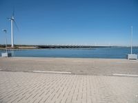 a brick street next to water under a blue sky with wind turbines in the distance