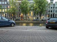 several cars parked near the canal and buildings in the evening with lights on a building in the background