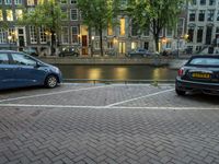 several cars parked near the canal and buildings in the evening with lights on a building in the background