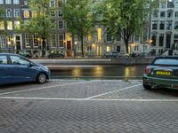 several cars parked near the canal and buildings in the evening with lights on a building in the background