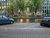 several cars parked near the canal and buildings in the evening with lights on a building in the background