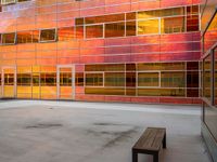 an empty bench sitting in front of a building with a lot of windows that has sun shining through the windows