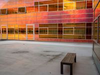 an empty bench sitting in front of a building with a lot of windows that has sun shining through the windows