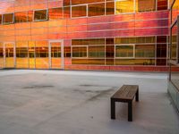 an empty bench sitting in front of a building with a lot of windows that has sun shining through the windows