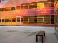 an empty bench sitting in front of a building with a lot of windows that has sun shining through the windows