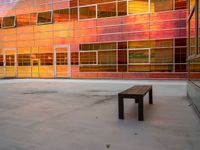 an empty bench sitting in front of a building with a lot of windows that has sun shining through the windows