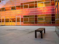 an empty bench sitting in front of a building with a lot of windows that has sun shining through the windows