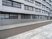 a paved sidewalk is by the street and building on one side there are windows on the building