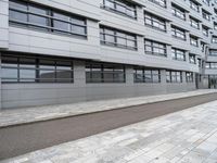 a paved sidewalk is by the street and building on one side there are windows on the building