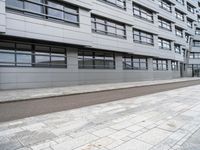 a paved sidewalk is by the street and building on one side there are windows on the building