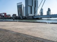 a brick walkway next to the water in front of buildings with cranes on them and some red boats