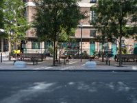 some park benches and trees on the street by a building that is next to a road