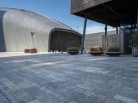 a brick patio with planters and a large metal building in the background, also two smaller cement planters