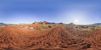 a red dirt track running across some dry plants in a canyon with bright sky above it