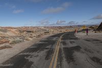 a woman walking down a empty street past rocks and shrubss while a man watches