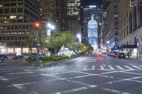 a city street that has lights on in the background, a fountain is in front of tall buildings and trees