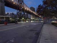 a paved road with buildings and bridge at night behind it in front of trees, in the distance,