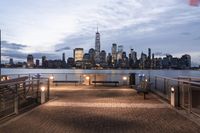 a long bench sits on the bridge overlooking a city skyline at dusk with buildings and lights