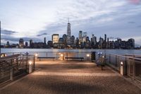 a long bench sits on the bridge overlooking a city skyline at dusk with buildings and lights