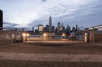 a long bench sits on the bridge overlooking a city skyline at dusk with buildings and lights