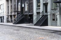 a person standing by some fire hydrants on a street near buildings and stairs and brick pavement