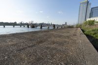 the view of a city skyline from an open river bank with a bridge and benches at the edge