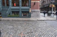 people walk by the sidewalk of a street corner, and look at a brick building in the background