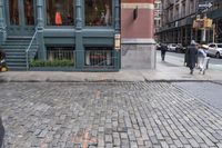 people walk by the sidewalk of a street corner, and look at a brick building in the background