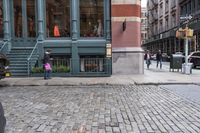 people walk by the sidewalk of a street corner, and look at a brick building in the background