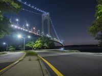 a night view of the bridge from an empty road near some water, and trees