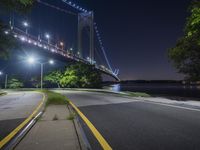 a night view of the bridge from an empty road near some water, and trees