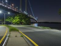 a night view of the bridge from an empty road near some water, and trees