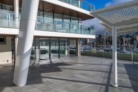 a large walkway leads to a building on the waterfront with windows open and view of sail boats