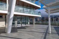 a large walkway leads to a building on the waterfront with windows open and view of sail boats